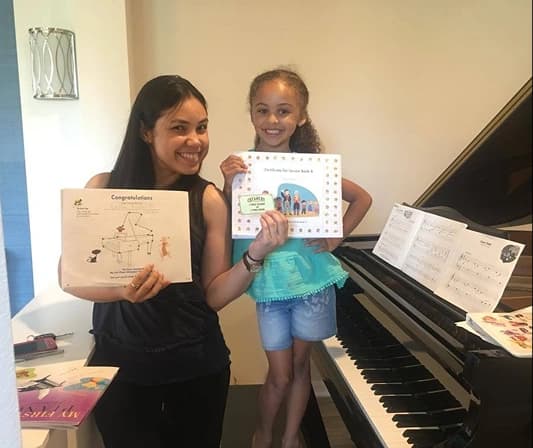 A smiling teacher and a young girl standing side by side next to a glossy black grand piano, each proudly holding up a certificate of achievement.