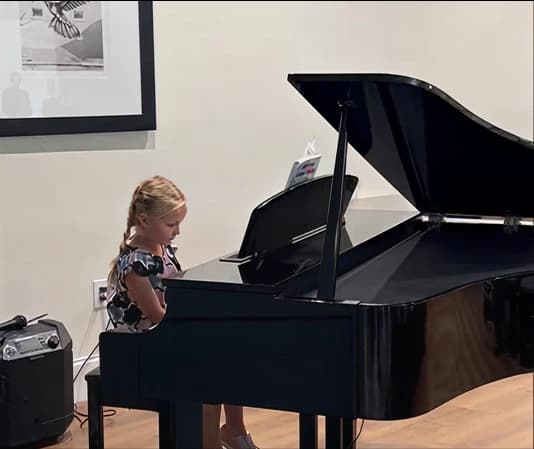 A young blonde student sits at a glossy black grand piano, intently reading the sheet music propped on the music stand.