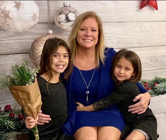 A smiling woman in a royal blue dress sits on a bench with her arms around two young girls in matching black dresses—one holding a brown-paper–wrapped bouquet—posed in front of a rustic wooden wall adorned with oversized silver and white holiday ornaments and greenery.